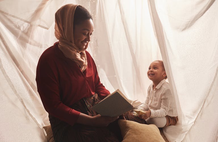A mother and child share a joyful moment reading together in a cozy indoor tent setting.