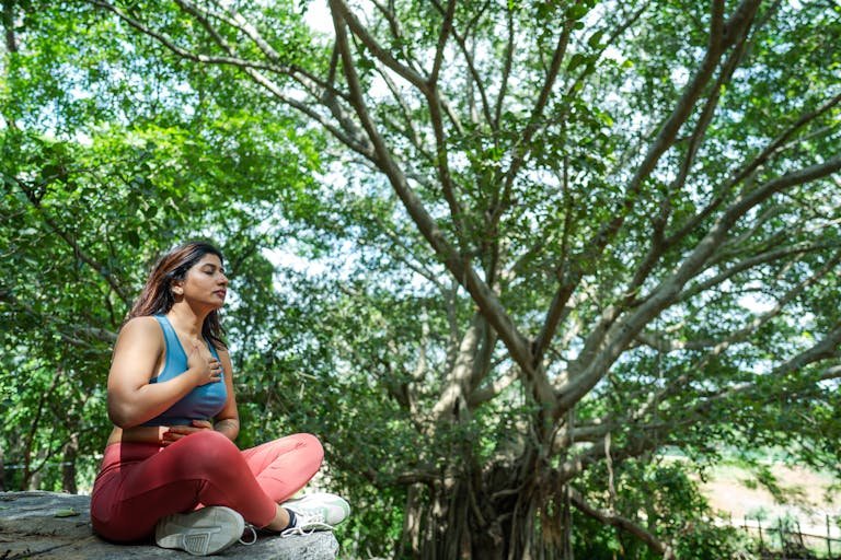 A woman meditates in a serene outdoor setting, surrounded by lush greenery.