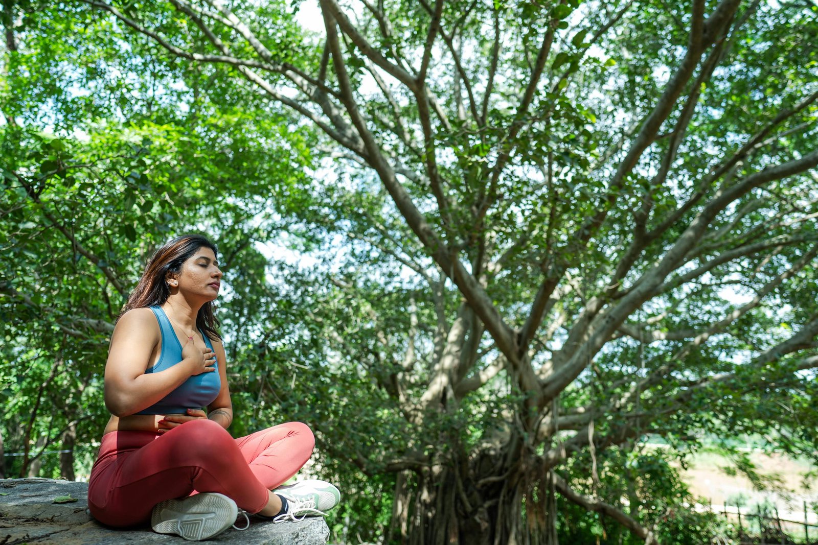 A woman meditates in a serene outdoor setting, surrounded by lush greenery.