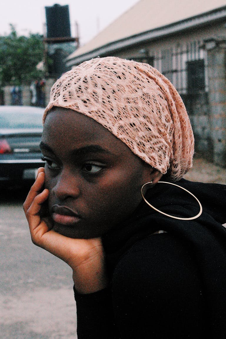 A pensive young woman wearing a headscarf rests her chin on her hand outdoors in Nigeria.