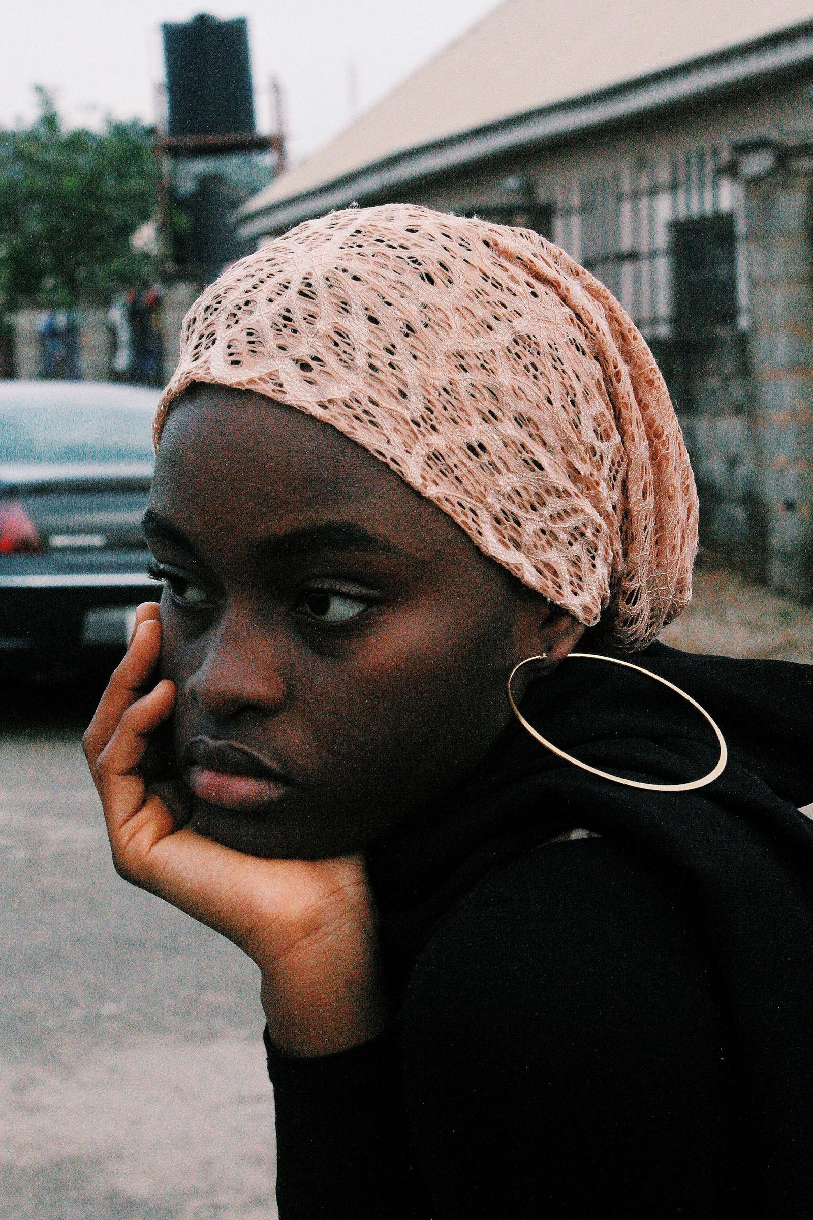 A pensive young woman wearing a headscarf rests her chin on her hand outdoors in Nigeria.
