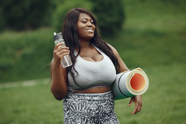 African American woman smiling with yoga mat and water bottle, ready for outdoor exercise.