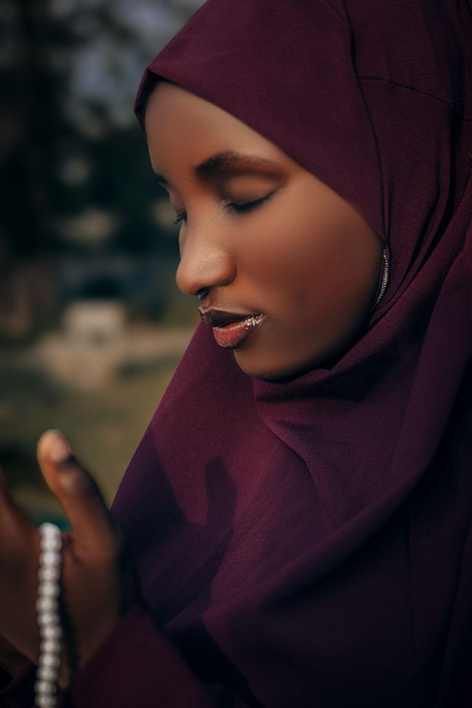 A contemplative Muslim woman wearing a maroon hijab, holding prayer beads while praying outdoors.