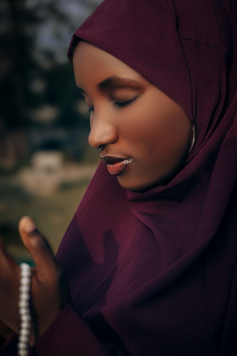 A contemplative Muslim woman wearing a maroon hijab, holding prayer beads while praying outdoors.