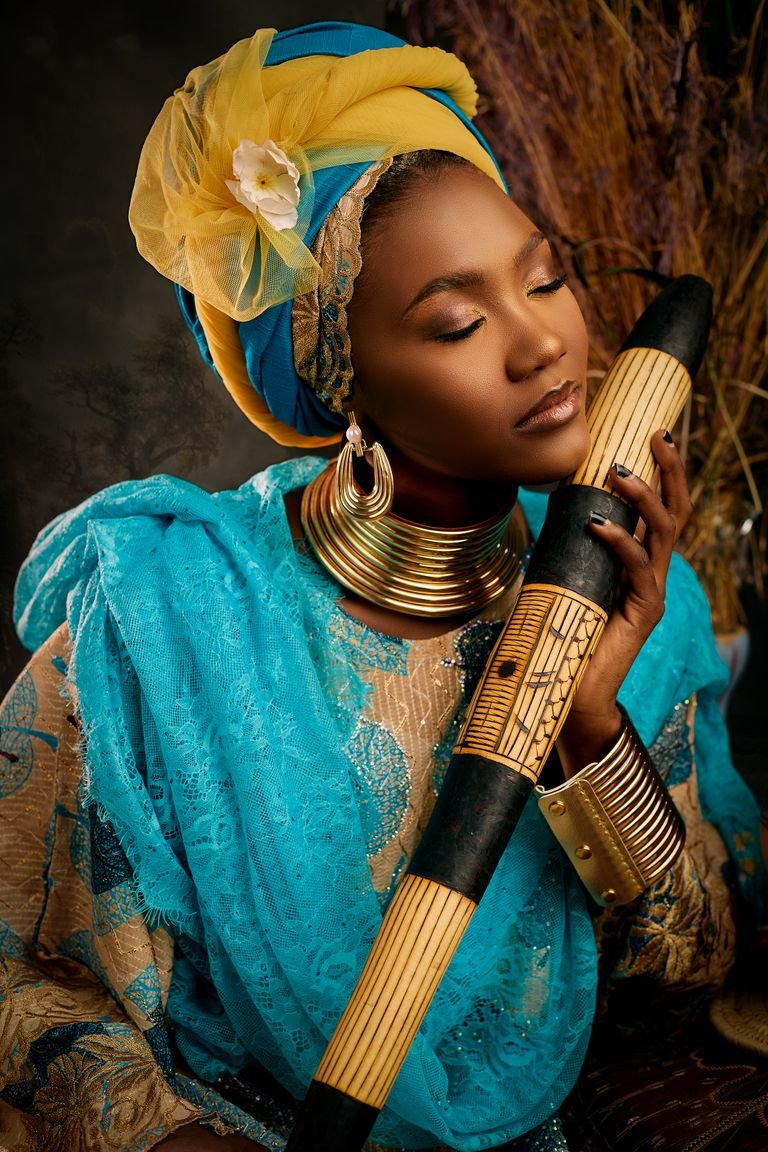 Elegant Nigerian woman poses with traditional instrument, showcasing cultural attire and jewelry.