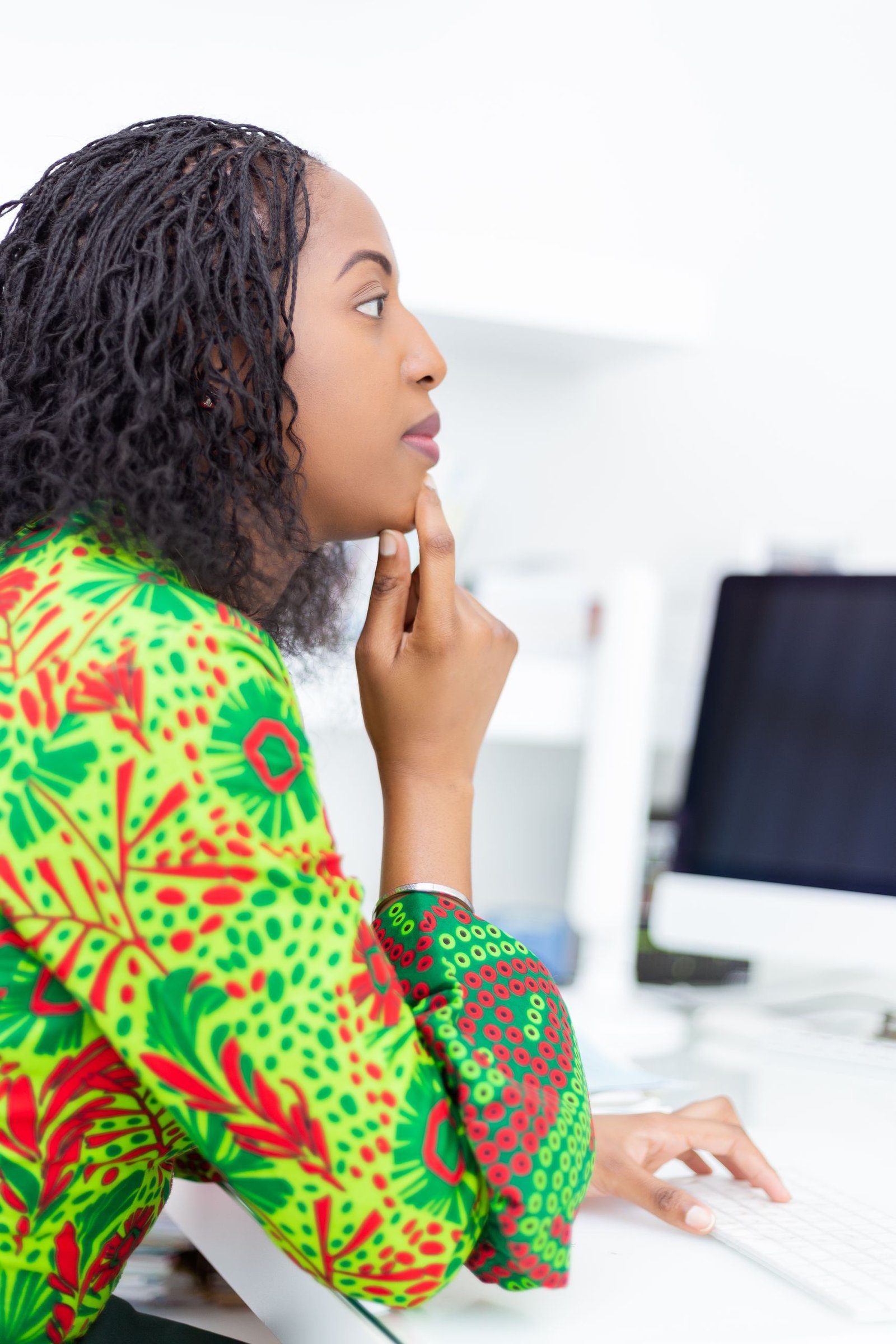 African woman in a bright green pattern dress, focused at her desk in an office setting.