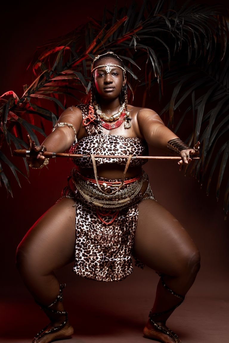 Plump mature African woman in traditional wear with accessories performing rite with wooden stick while standing with reached arms and looking at camera in darkness