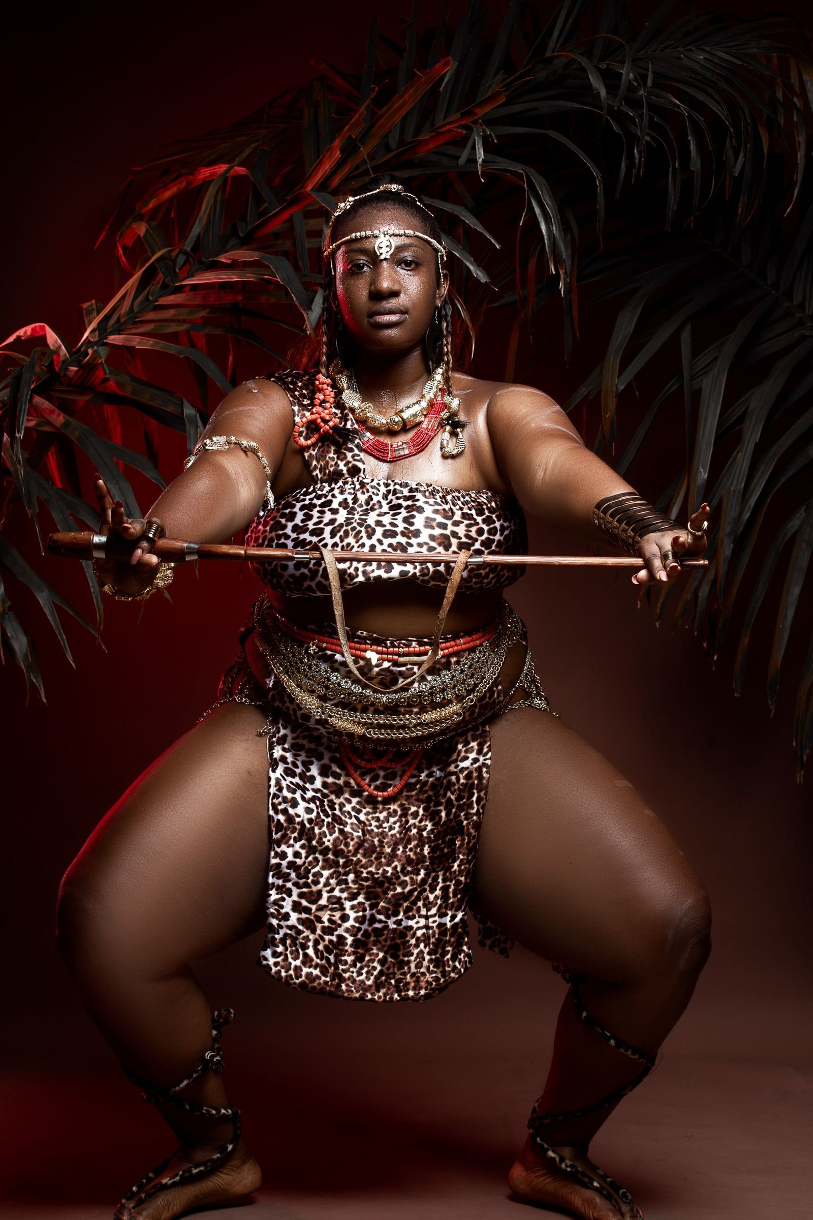 Plump mature African woman in traditional wear with accessories performing rite with wooden stick while standing with reached arms and looking at camera in darkness