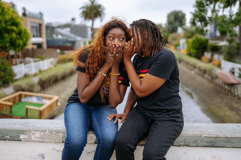Two women share a secret while sitting outdoors on a ledge.